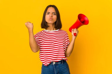 Young hispanic woman holding a megaphone showing fist to camera, aggressive facial expression. clipart