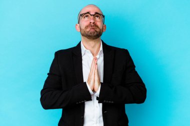 Genç, beyaz, kel bir adam mavi arka planda izole edilmiş, el ele tutuşmuş, ağzı yakınken dua ediyor, kendinden emin..