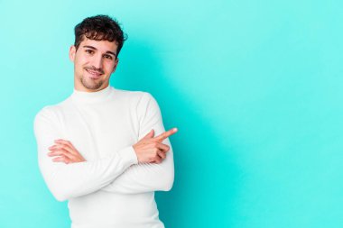 Mavi arka planda izole edilmiş genç beyaz adam işaret parmağıyla işaret ederek gülümsüyor..