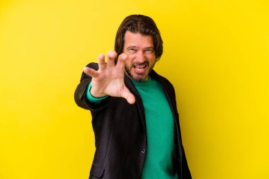 Sarı arka planda izole edilmiş orta yaşlı beyaz adam pençelerini kedi taklidi yaparken gösteriyor, agresif bir hareket..