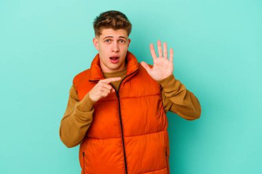 Mavi arka planda izole edilmiş genç beyaz adam gülümseyerek beş numarayı parmaklarıyla gösteriyor..