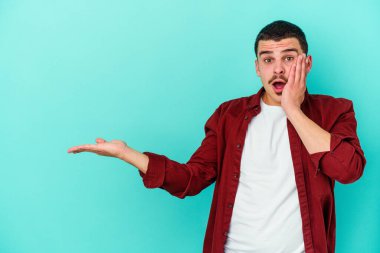 Mavi arka planda izole edilmiş genç beyaz adam, avuç içi fotokopi alanını elinde tutmaktan etkilenmiş..