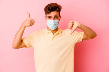 Koronavirüs için koruyucu takan beyaz bir adam pembe arka planda izole edilmiş, baş parmağını kaldırıyor, zor bir seçim.