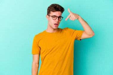 Mavi arka planda izole edilmiş genç beyaz adam bir gömlek fotokopi alanını işaret ediyor, gururlu ve kendinden emin.