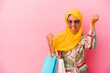 Genç Müslüman kadın pembe arka planda izole giysiler alıyor zafer kazandıktan sonra yumruğunu kaldırıyor, kazanan konsept..