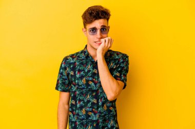 Hawaii gömleği giyen genç beyaz adam sarı arka planda izole edilmiş tırnak yiyor, gergin ve endişeli..