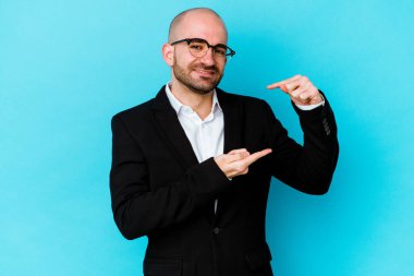Genç, beyaz, kel bir adam mavi arka planda izole edilmiş. Elinde parmakları olan, gülümseyen ve kendinden emin bir şey var..
