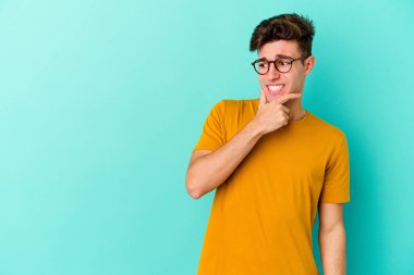 Kafkasyalı genç adam mavi arka planda izole edilmiş kafasının arkasına dokunuyor, düşünüyor ve bir seçim yapıyor..