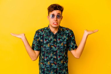 Sarı arka planda izole edilmiş Hawaii tişörtü giyen genç beyaz bir adam. Kopya alanını tutmak için kafası karışık ve omuz silkme konusunda şüpheci..