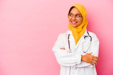 Genç doktor müslüman kadın pembe arka planda izole edilmiş. Kollarını kavuşturarak gülümsüyor..