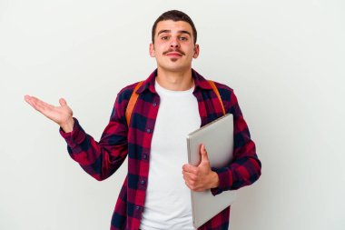 Beyaz arka planda dizüstü bilgisayar tutan beyaz tenli genç bir öğrenci kollarıyla ölçek oluşturur, mutlu ve kendinden emin hisseder..