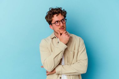 Gözlüklü genç beyaz adam mavi arka planda izole edilmiş düşünüyor ve yukarı bakıyor, yansıtıyor, düşünüyor, hayal kuruyor..