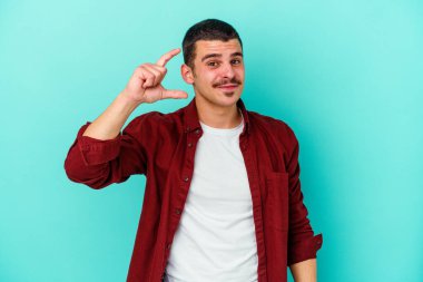 Kafkasyalı genç adam mavi arka planda izole edilmiş. Elinde işaret parmaklarıyla küçük bir şey tutuyor, gülümsüyor ve kendinden emin..
