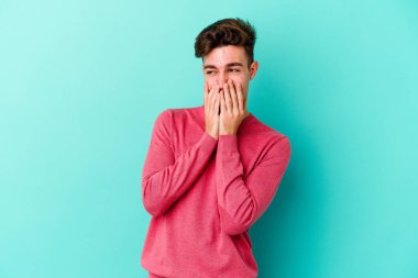 Mavi arka planda izole edilmiş genç beyaz bir adam bir şeye gülüyor, ağızlarını ellerle kapatıyor..