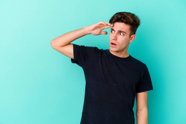 Kafkasyalı genç adam mavi arka planda izole edilmiş, uzaklara bakıyor ve elini alnına koyuyor..