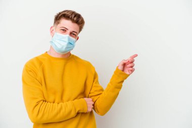 Koronavirüs için maske takan genç adam beyaz arka planda izole edilmiş, mutlu bir şekilde işaret parmağıyla işaret ederek gülümsüyor..