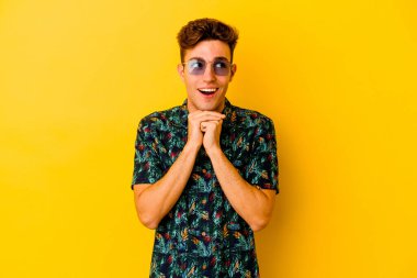Sarı arka planda izole edilmiş Hawaii gömleği giyen genç beyaz adam ellerini çenesinin altında tutuyor ve mutlu bir şekilde yan tarafa bakıyor..