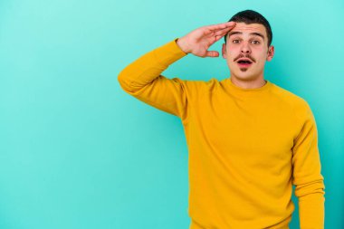 Kafkasyalı genç adam mavi arka planda izole edilmiş, uzaklara bakıyor ve elini alnına koyuyor..