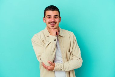 Mavi arka planda izole edilmiş genç beyaz adam gülümsüyor ve kendinden emin, elleriyle çenesine dokunuyor..