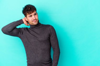 Kafkasyalı genç adam mavi arka planda izole edilmiş kafasının arkasına dokunuyor, düşünüyor ve bir seçim yapıyor..