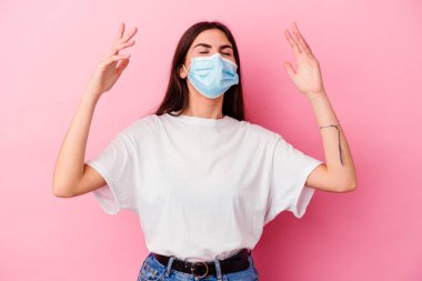 Virüs maskesi takan beyaz bir kadın pembe arka planda izole edilmiş, gökyüzüne haykırıyor, yukarı bakıyor, hayal kırıklığına uğruyor..