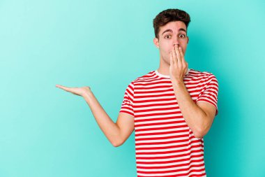 Mavi arka planda izole edilmiş genç beyaz adam, avuç içi fotokopi alanını elinde tutmaktan etkilenmiş..