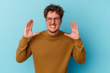 Mavi arka planda izole edilmiş gözlüklü genç beyaz adam öfkeyle bağırıyor..