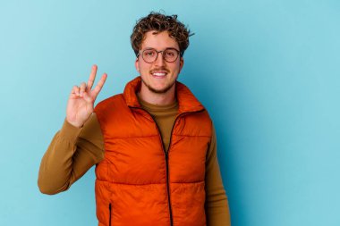 Mavi arka planda gözlüklü genç beyaz adam zafer işareti gösteriyor ve gülümsüyor..