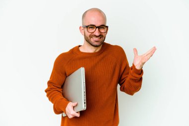 Beyaz arka planda dizüstü bilgisayar tutan genç kel bir adam hoş bir sürprizle karşılaşıyor, heyecanlı ve el kaldırıyor..