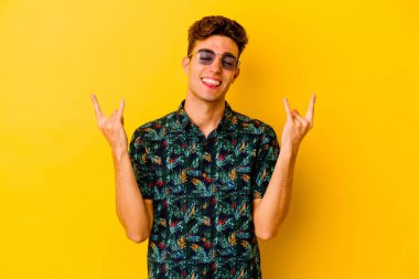 Sarı arka planda izole edilmiş Hawaii tişörtü giyen genç beyaz adam parmaklarıyla kaya hareketi gösteriyor.