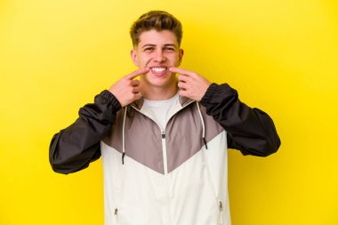 Sarı arka planda izole edilmiş genç beyaz adam gülümsüyor, parmaklarını ağzına doğrultuyor..