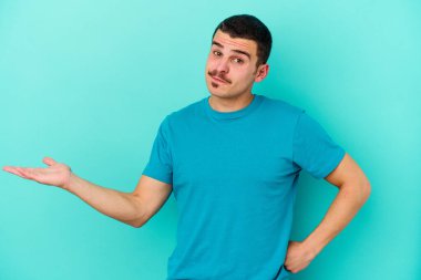 Kafkasyalı genç adam mavi arka planda izole edilmiş sorgulama jestinde şüphe ediyor ve omuz silkiyor..