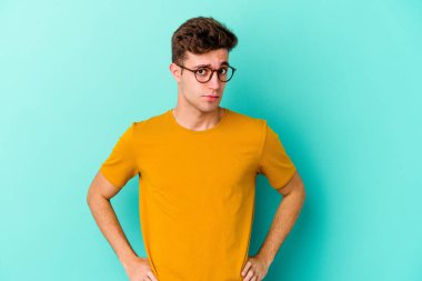 Kafkasyalı genç adam mavi arka planda izole, kafası karışmış, şüpheli ve güvensiz hissediyor..