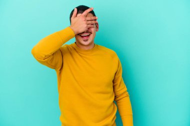 Mavi arka planda soyutlanmış genç beyaz bir adam elleriyle gözlerini kapatıyor, geniş bir gülümsemeyle bir sürpriz bekliyor..