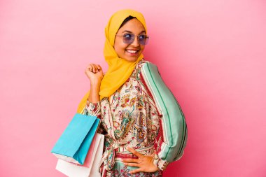 Genç Müslüman kadın pembe arka planda izole giysiler alıyor. Gülümsüyor, neşeli ve keyifli görünüyor..