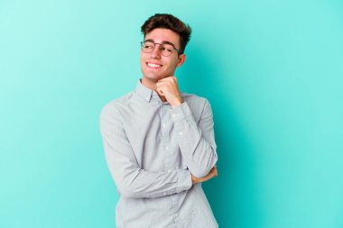 Mavi arka planda izole edilmiş genç beyaz adam gülümsüyor ve kendinden emin, elleriyle çenesine dokunuyor..