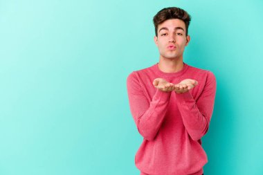 Kafkasyalı genç adam mavi arka planda izole edilmiş dudaklar kıvrılıyor ve hava öpücüğü göndermek için avuç içleri tutuyor..