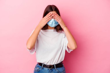 Virüs maskesi takan beyaz bir kadın pembe arka planda izole edilmiş gözleri ellerle kaplıyor, büyük bir gülümsemeyle sürpriz bekliyor..