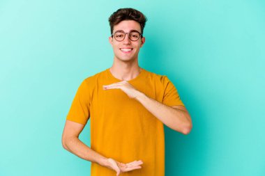 Mavi arka planda izole genç beyaz adam iki eliyle bir şey tutuyor, ürün sunumu..