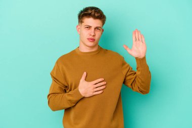 Kafkasyalı genç adam mavi arka planda izole edilmiş yemin ediyor, elini göğsüne koyuyor..