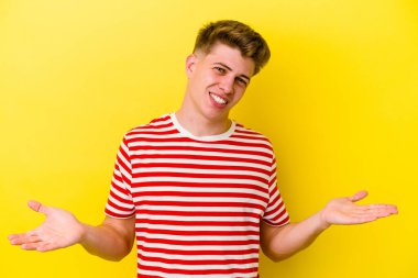 Sarı arka planda izole genç beyaz adam kollarıyla ölçek yapar, mutlu ve kendinden emin hisseder..