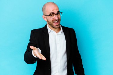 Genç, beyaz, kel bir adam mavi arka planda izole edilmiş. Kameraya doğru elini uzatarak selam veriyor..