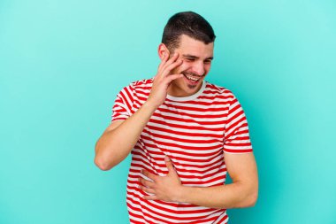 Mavi arka planda izole edilmiş genç beyaz adam mutlu bir şekilde gülüyor ve ellerini karnının üstünde tutarak eğleniyor..