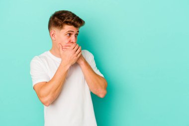 Kafkasyalı genç adam mavi arka planda izole edilmiş. Düşünceli bir şekilde ağzıyla ağzını kapatan bir fotokopi odasına bakıyor..