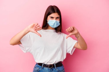 Virüs maskesi takan beyaz bir kadın pembe arka planda izole edilmiş, baş parmağını aşağı gösteren ve hoşlanmadığını ifade eden..