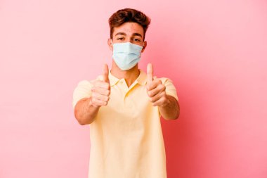 Koronavirüs için koruyucu takan beyaz bir adam pembe arka planda izole edilmiş, baş parmakları olan, bir konuda tezahürat yapan, destek ve saygı duyulan bir konsept..