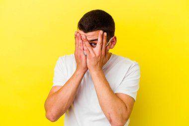 Koronavirüs için koruyucu takan beyaz bir adam sarı arka planda izole edilmiş. Korkmuş ve gergin parmaklarıyla göz kırp..