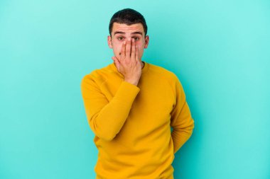 Mavi arka planda izole edilmiş genç beyaz adam şok olmuş, ağzı ellerle kaplanmış, yeni bir şey keşfetmeye hevesli..