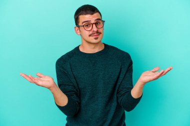 Kafkasyalı genç adam mavi arka planda izole edilmiş sorgulama jestinde şüphe ediyor ve omuz silkiyor..