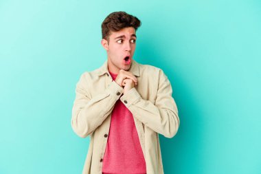 Kafkasyalı genç adam mavi arka planda izole edilmiş şans için dua ediyor, şaşırmış ve ağzını açıp önüne bakıyor..